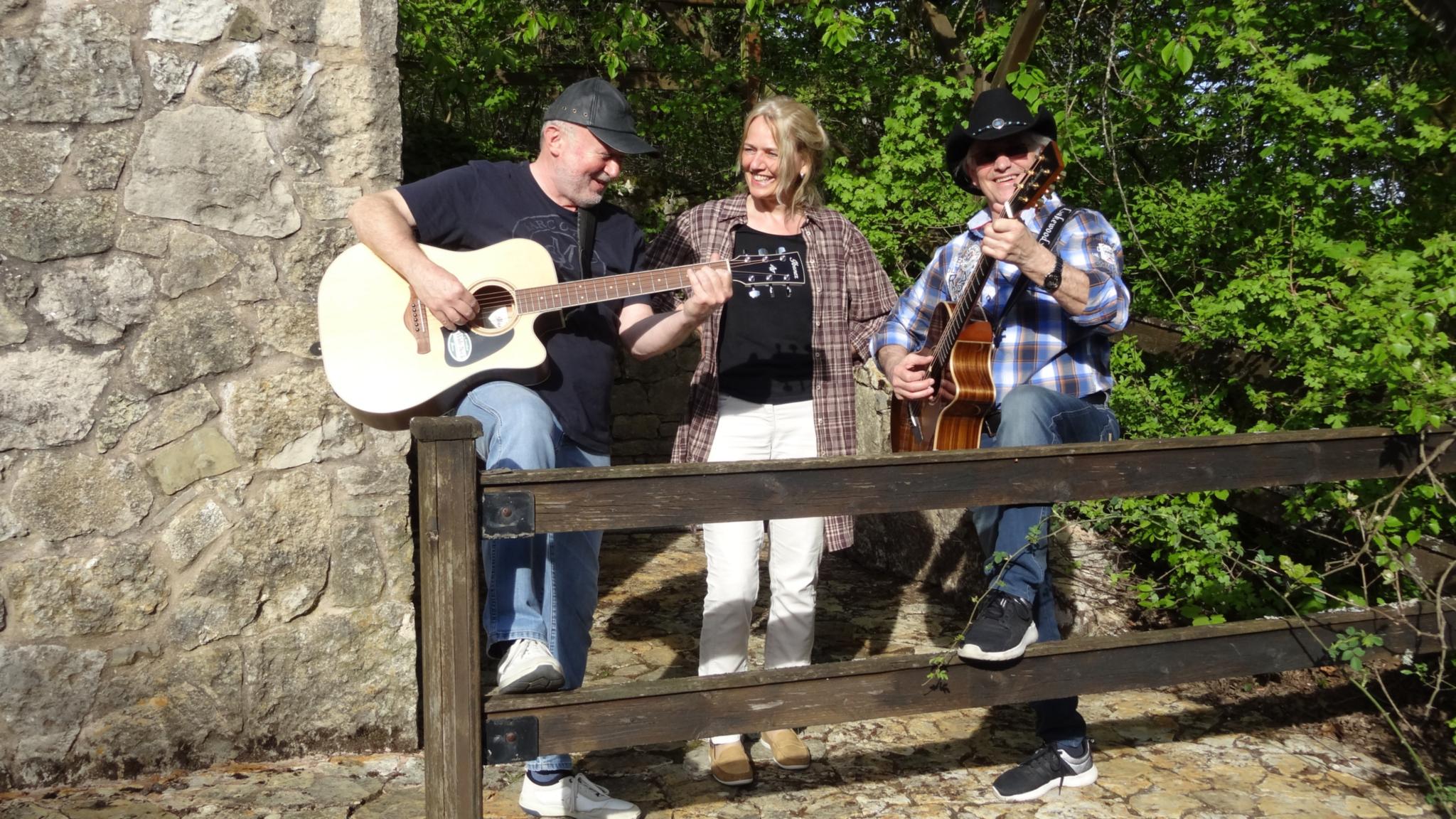 Zwei Männer mit Gitarre und eine Frau stehen neben einer groben Steinwand. Vor ihnen sind die Balken eines Zauns zu sehen. Auf der rechten Bildseite sind grüne Büsche.