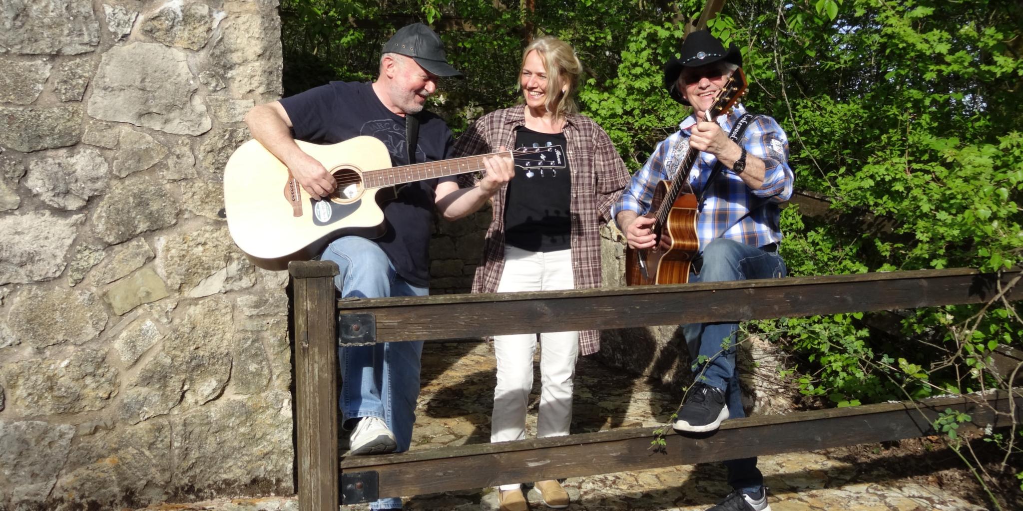 Zwei Männer mit Gitarre und eine Frau stehen neben einer groben Steinwand. Vor ihnen sind die Balken eines Zauns zu sehen. Auf der rechten Bildseite sind grüne Büsche. (c) Steely Tones Zwei Männer mit Gitarre und eine Frau stehen neben einer groben Steinwand. Vor ihnen sind die Balken eines Zauns zu sehen. Auf der rechten Bildseite sind grüne Büsche.