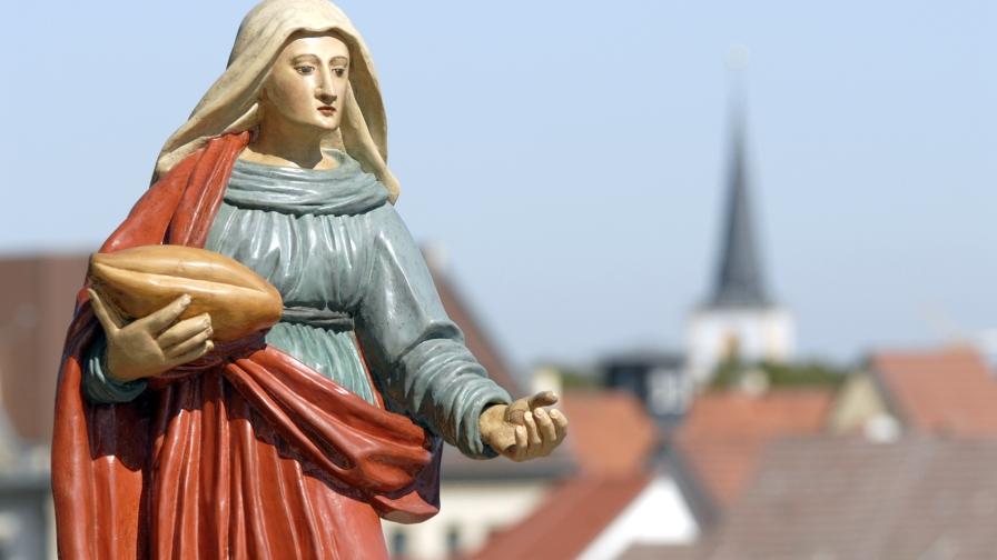 Eine Frauenfigur mit einem Brot in der Hand, im Hintergrund Hausdächer.