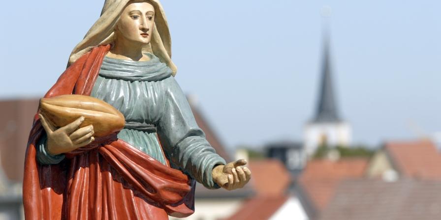 Eine Frauenfigur mit einem Brot in der Hand, im Hintergrund Hausdächer.